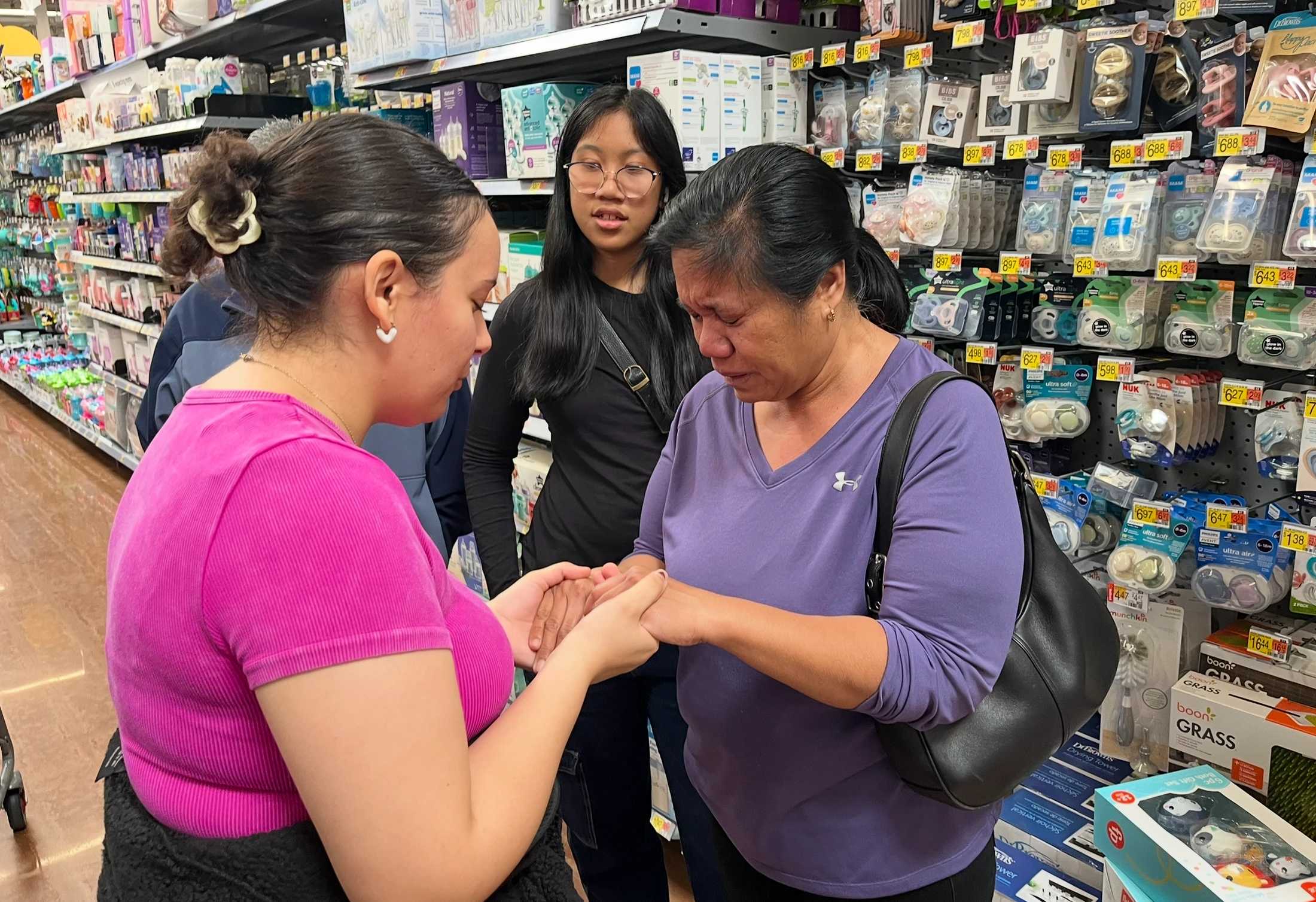 A woman in a pink shirt comforts an emotional woman in a purple shirt by holding her hands in a store's baby aisle as a younger woman looks on.